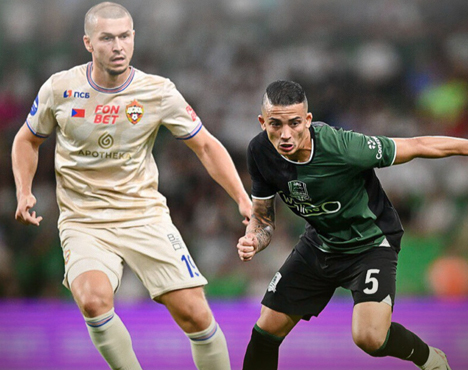 Два футболиста в игровом моменте на поле: спортсмен в бежевой форме справа против игрока в черной форме слева. Динамичная обстановка с размытым фоном зрителей и стадиона