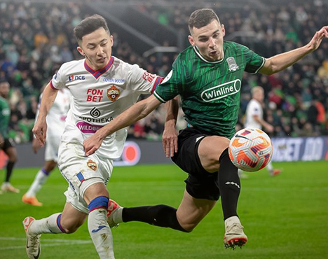 Футболист в зелёной форме пытается обыграть соперника в белой форме на переполненном стадионе. Игроки сосредоточены на мяч, который летит в воздухе. На заднем плане видны размытые фигуры зрителей