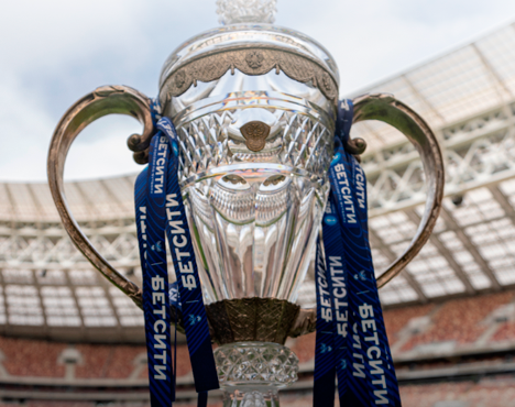 Серебряный кубок с ручками и синими лентами, украшенный узорами, на фоне стадиона