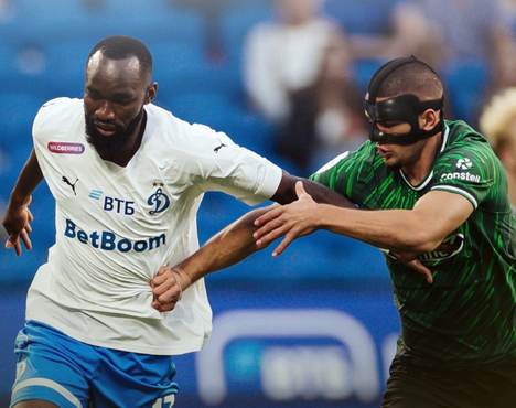 На изображении два футболиста в борьбе за мяч. Игрок в белой форме с логотипом BetBoom и Динамо противостоит сопернику в зелёно-чёрной форме, который носит чёрную маску на лице, символизирующую защиту