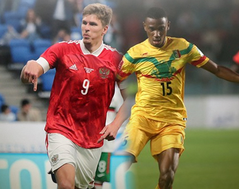 На изображении два футболиста в момент игры: один в красной форме сборной России, второй в желтой форме с зелеными и красными полосами, представляющей сборную Мали. Они находятся на фоне футбольного стадиона