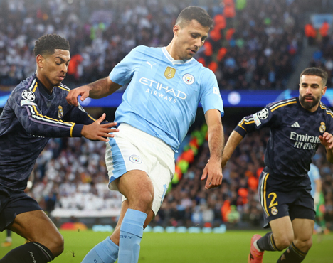 Футбольный матч: игрок в голубой форме с мячом, соперники в тёмной форме пытаются его обыграть. Напряжённый игровой момент, трибуны заполнены зрителями