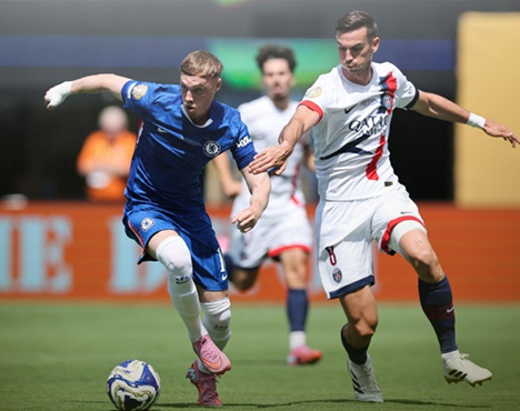 На изображении два футболиста борются за мяч во время матча. Один из игроков в синей форме, другой в белой с красно-синими элементами. Футболисты активно передвигаются по полю. На заднем плане видны размыленные фигуры людей