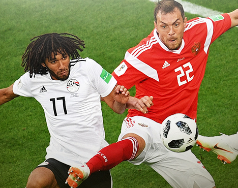 Два футболиста в динамичной борьбе за мяч во время матча. Спортсмен в красной форме сражается с игроком в белой, оба стремятся к овладению футбольным мячом на поле